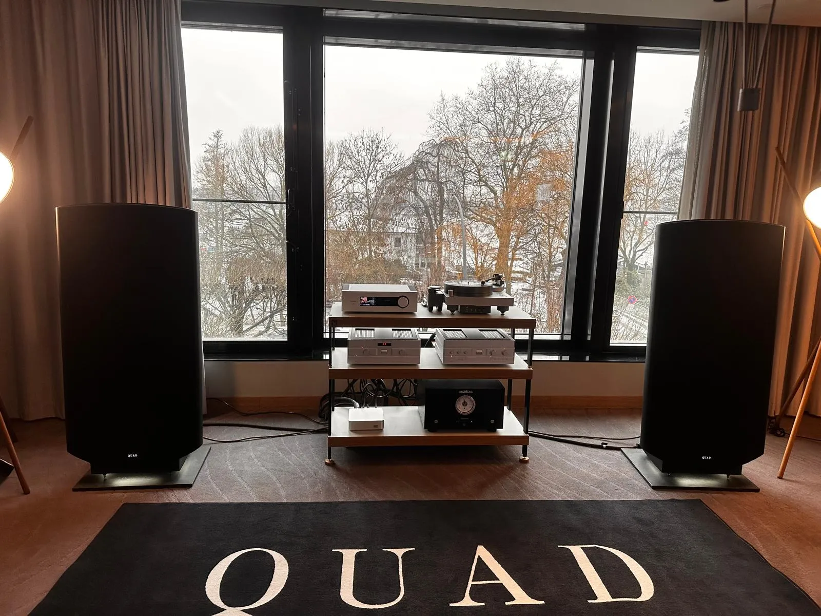 Eine Quad-Anlage: zwei Standlautsprecher links und rechts, dazwischen ein Rack mit Verstärkern, Plattenspieler und Netzteil, davor ein Teppich mit dem Quad-Schriftzug.