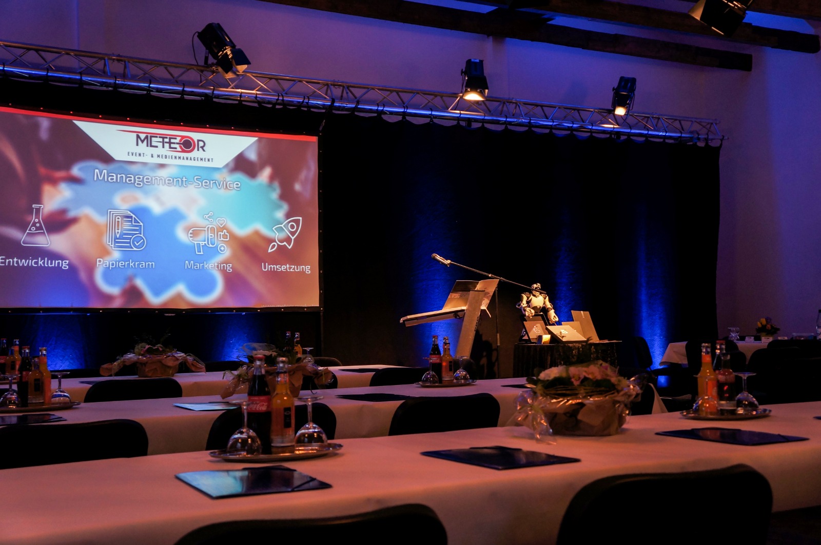 Die Bühne im Saal: eine Leinwand mit einer Präsentationsfolie, darüber eine Traverse mit Scheinwerfern, davor weiß eingedeckte Tafeln in blauem Licht.